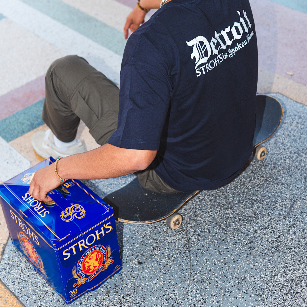 Person sitting on a skateboard holding a box of Stroh's beer, wearing a 'Detroit' Stroh's t-shirt.